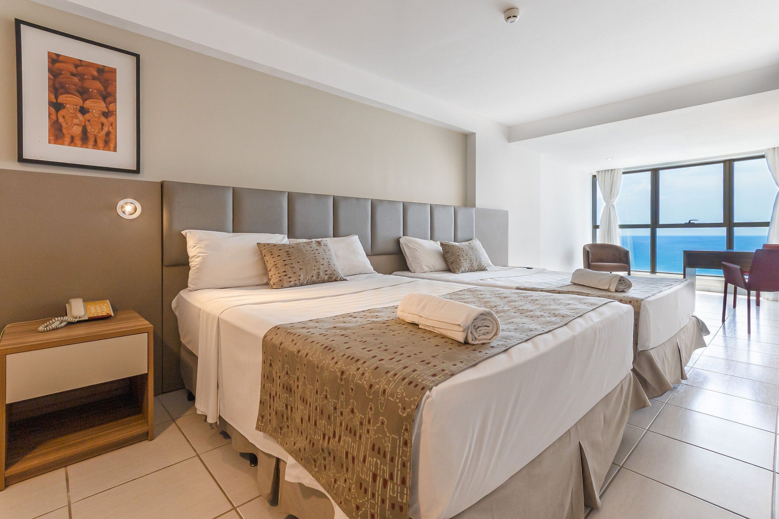 Habitación de hotel con dos camas, una mesita de noche, una silla y un ventanal con vistas.