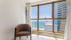 A room with a large window, a brown chair, and a view of the sea and buildings. - Beach Class #1104 - Quadruplo por Carpediem (Recife)