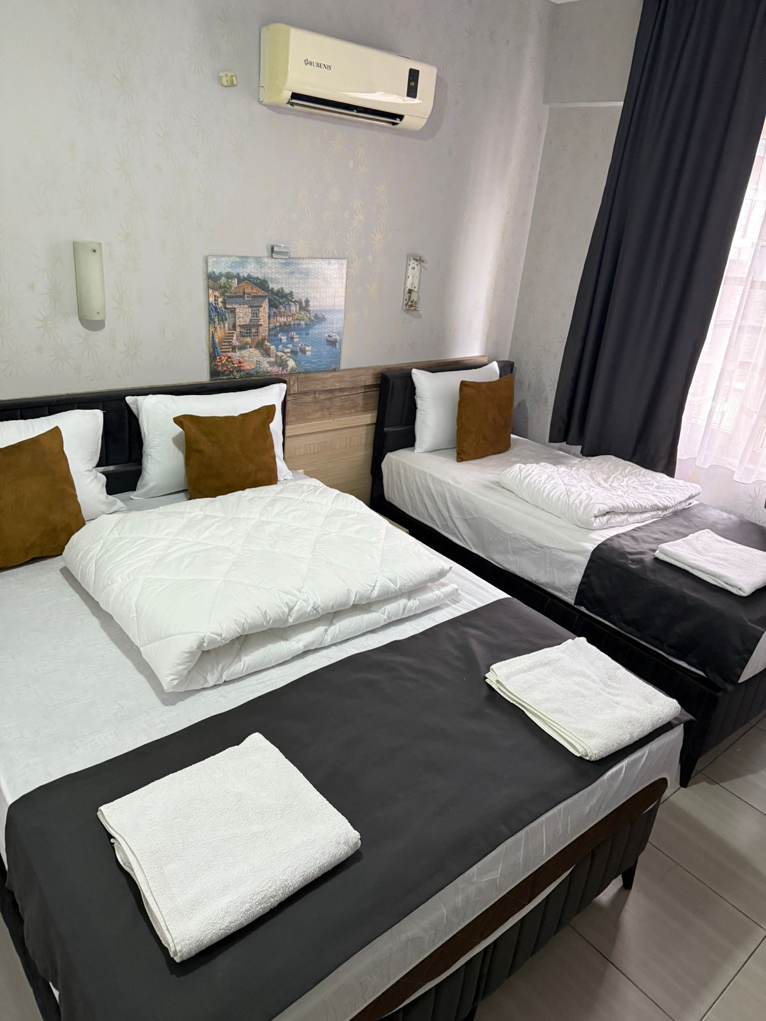 A hotel room with two beds, a painting on the wall, an air conditioner, and a window with curtains.
