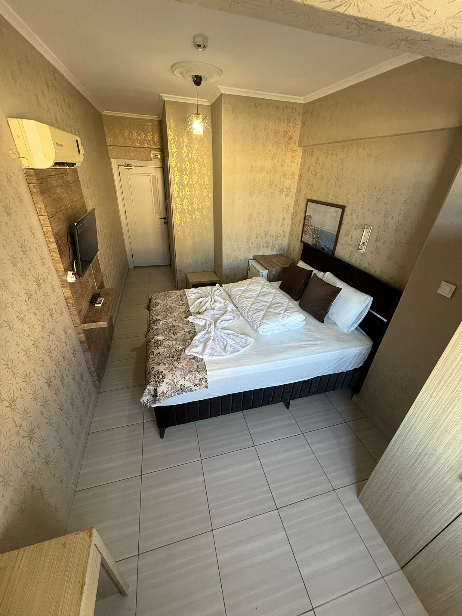 Una habitación de hotel compacta con una cama, una mesita de noche pequeña, un televisor montado en la pared y una puerta que da a un baño.