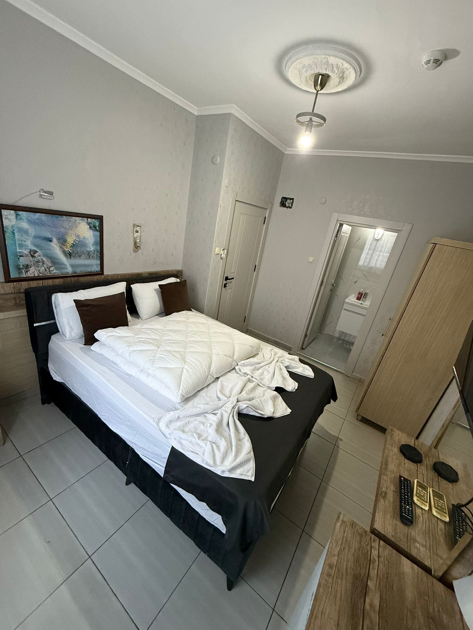 A neatly made bed with white and brown bedding, a wooden nightstand with remote controls, a wooden wardrobe, and a painting on the wall.