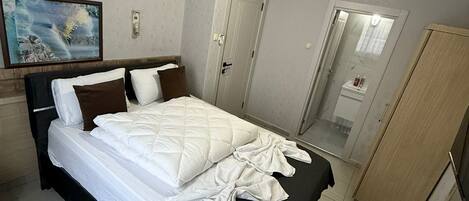 A neatly made bed with white and brown bedding, a wooden nightstand with remote controls, a wooden wardrobe, and a painting on the wall.
