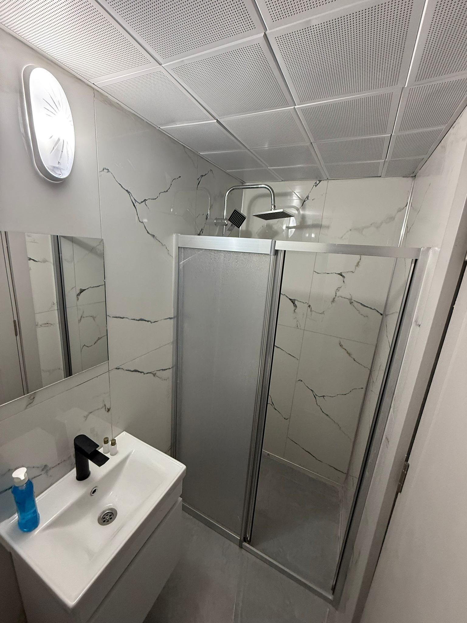 A modern bathroom with a white sink, a mirror, a shower enclosure, and a ceiling with a perforated design.