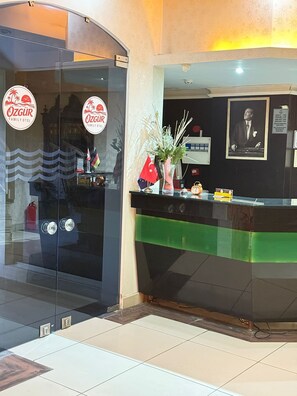 Zona de recepción de un hotel con puerta de vidrio, mostrador de recepción, un cuadro enmarcado en la pared y un cartel.