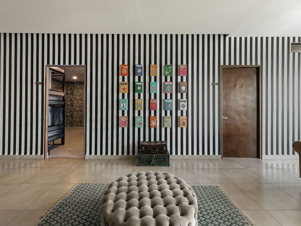 Une pièce avec un mur à rayures, un pouf rond et une valise posée sur un tapis.