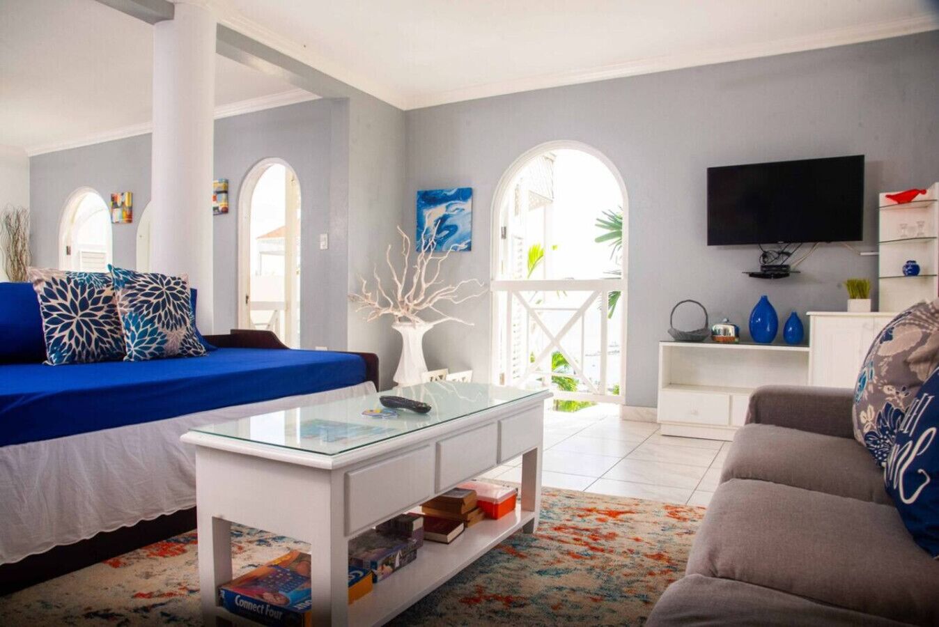 A modern living room with a blue sofa, a glass coffee table, and a grey couch. There is a flat-screen TV mounted on the wall and a bookshelf with various items.