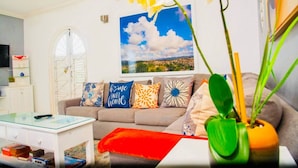A living room with a sofa, a coffee table, and a bookshelf. There is a painting on the wall and a window with shutters.