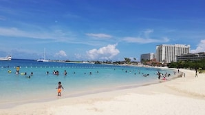 A beach with clear blue water, people swimming and walking on the sand, and a large hotel in the background.