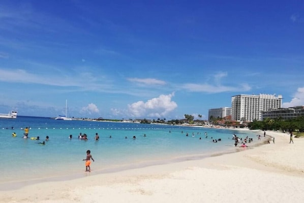 Uma praia com água azul cristalina, pessoas nadando e caminhando na areia, e um grande hotel ao fundo.