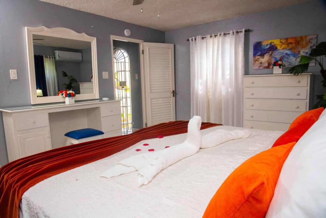 Een net opgemaakt bed met een witte deken en oranje kussens, een witte dressoir met lades, een blauwe kruk, een grote spiegel en een schilderij aan de muur.