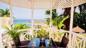 A covered outdoor seating area with wicker furniture and potted plants, overlooking a scenic ocean view.