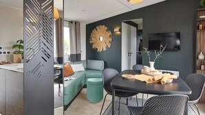 A modern living room with a sectional sofa, a dining table with chairs, a TV mounted on the wall, and a decorative wall piece. - ZEcamping 4* animé espace aquatique sauna hammam locatif TV LV BBQ (Aubigny-Les Clouzeaux)