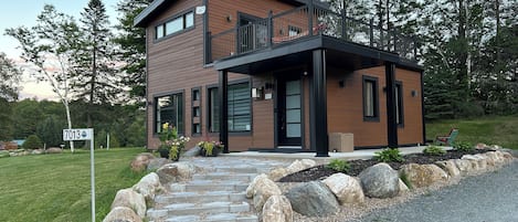 Une maison moderne à deux étages, dotée d’un escalier en pierre menant à l’entrée, et entourée d’un paysage naturel.