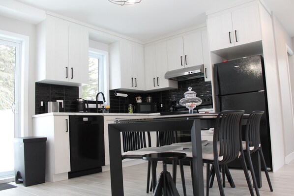 A modern kitchen with white cabinets, a black refrigerator, and a dining area with black chairs. - La Casa TiMoris (Wentworth-Nord)