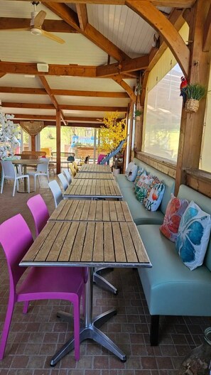 Een overdekt terras met houten tafels en stoelen, een gewatteerde bank en uitzicht op de omgeving.