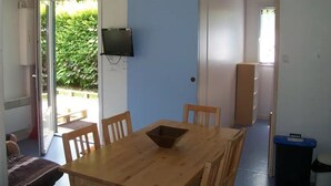 Quarto com mesa de madeira, cadeiras, TV fixada na parede e porta que dá para uma varanda com vegetação.