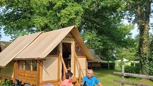 A tent with a wooden frame and a canvas roof, a bicycle, and a person sitting at a table. - ZEcamping 3* piscine locatif 59m2 4 pers (Montaut)