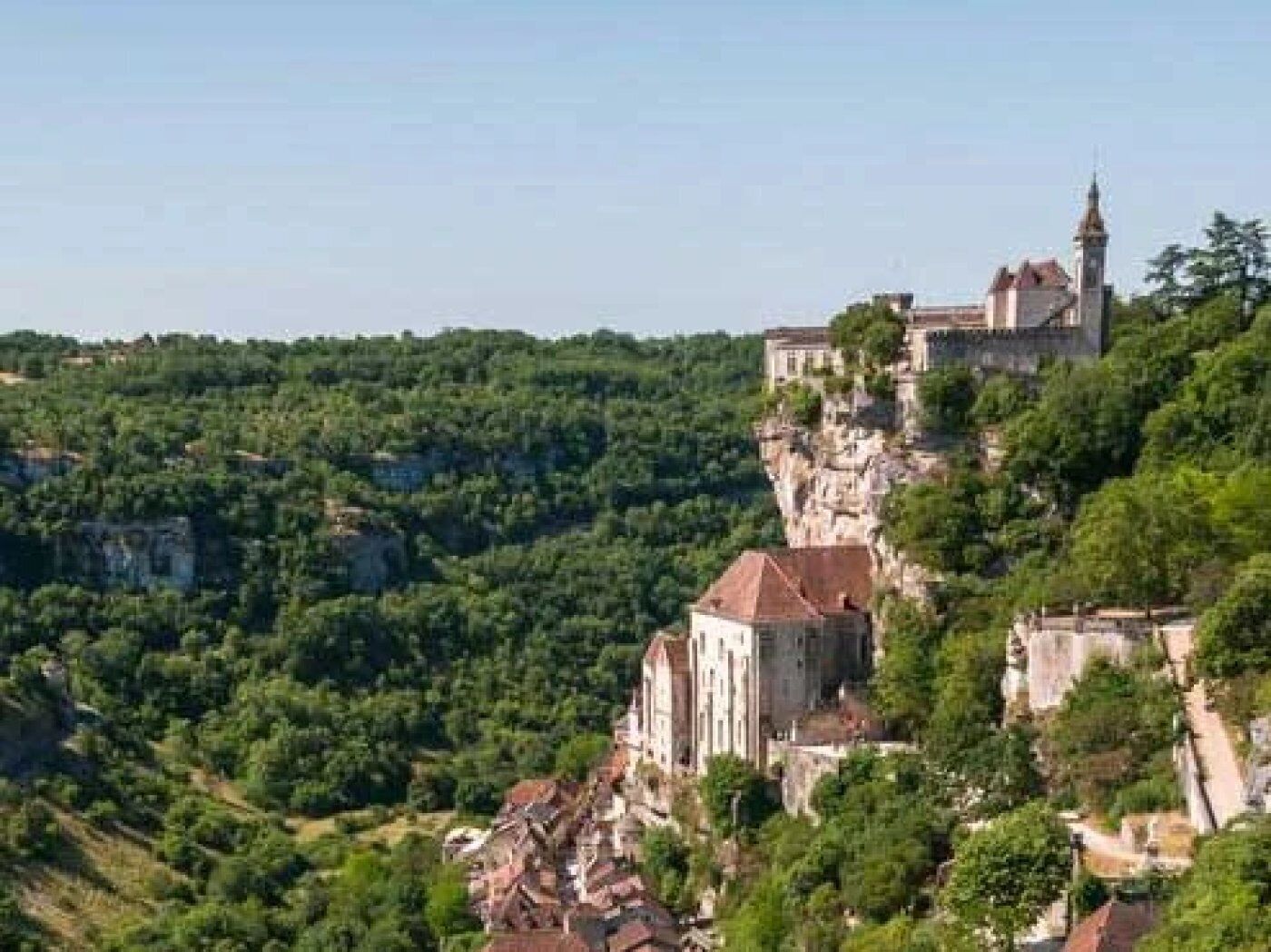 A historic building perched on a cliff with a church tower, surrounded by lush greenery.