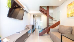 A modern living room with a flat-screen TV mounted on the wall, a sofa, a coffee table, and a staircase with glass railings. - Villa Del Porto #307 - Flat in Porto by Carpediem (Ipojuca)