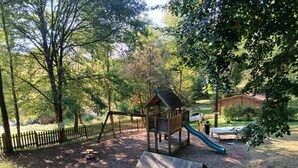Une aire de jeux avec un toboggan et une structure en bois, entourée d’arbres et délimitée par une clôture.