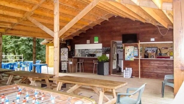 An outdoor wooden structure with tables and chairs, a foosball table, and a kitchen area.