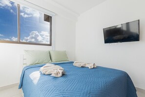A bedroom with a large window, a bed with blue bedding, two pillows, and a flat-screen TV on the wall. - Cupe Beach Living #102d - Triple by Carpediem (Ipojuca)