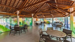 Um espaço interno com mesas e cadeiras, teto alto com vigas de madeira e grandes janelas com vidro esverdeado.