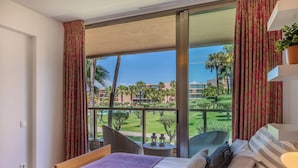 A hotel room with a large balcony, a bed with pillows, a small table with a lamp, and a view of a landscaped area with palm trees and buildings. - Salgados Resort Beach & Nature Escape (Albufeira)