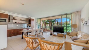 A modern kitchen with a dining area, a living room with a TV, and a large window with a view of palm trees. - Salgados Resort Beach & Nature Escape (Albufeira)