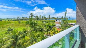 A balcony with a clear view of a lush green landscape, palm trees, and a distant building. - Cupe Garden #315 - Porto de Galinhas by Carpediem (Ipojuca)