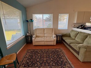 Un salon avec un canapé beige, un tapis à motifs et une fenêtre équipée de stores.