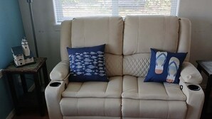 Un sofá reclinable color beige con dos almohadas estampadas, una mesita con una maqueta de una casa y una lámpara de pie.