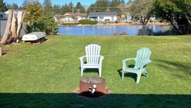 Un patio trasero con césped, dos sillas Adirondack, un brasero y una vista del lago y casas al fondo.