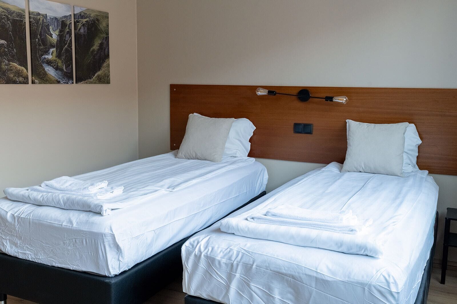 Deux lits simples avec du linge de lit blanc, une tête de lit en bois et un luminaire fixé au mur.