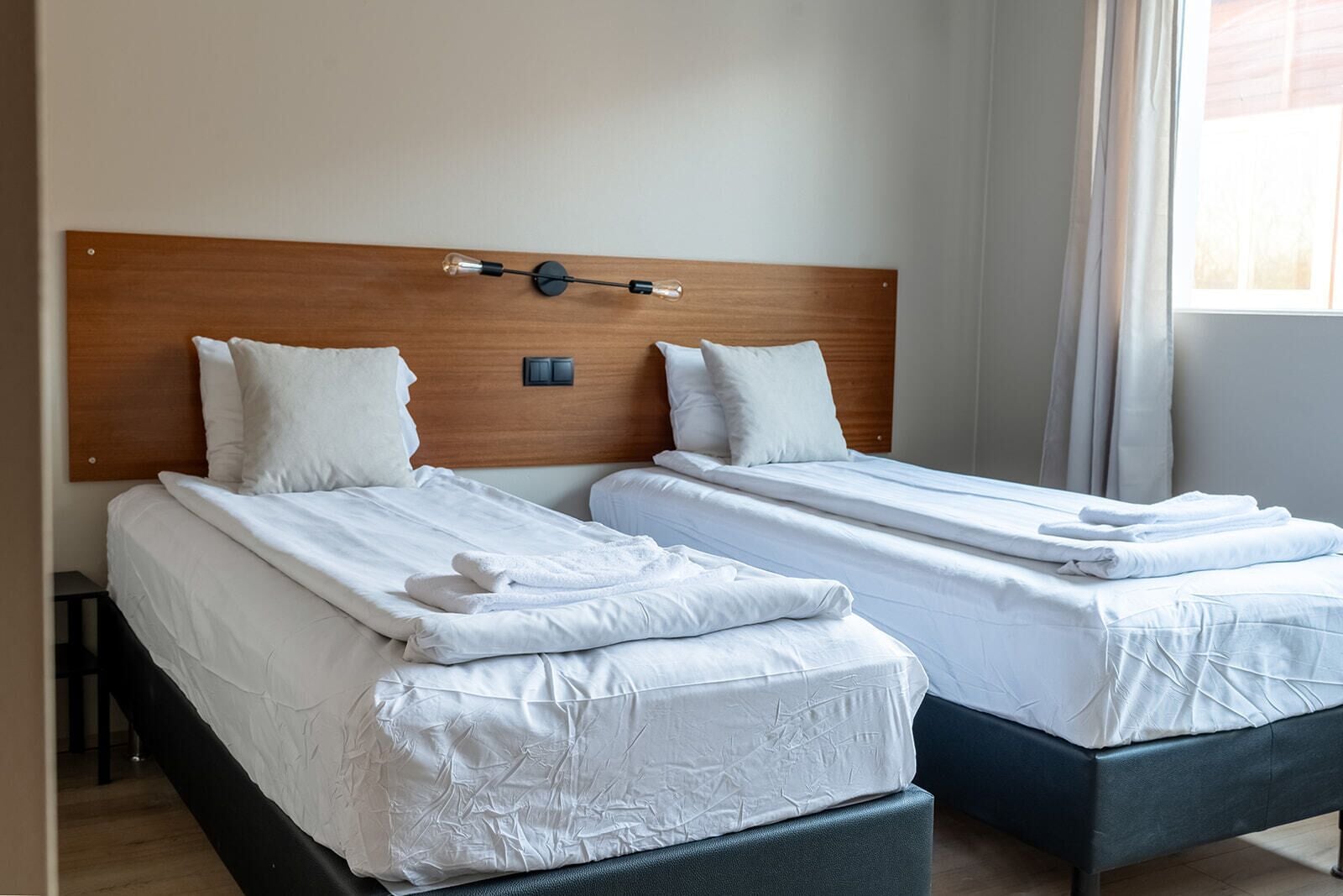 A hotel room with two single beds, each with white linens and pillows, a wooden headboard, and a bedside table.