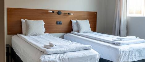 A hotel room with two single beds, each with white linens and pillows, a wooden headboard, and a bedside table.