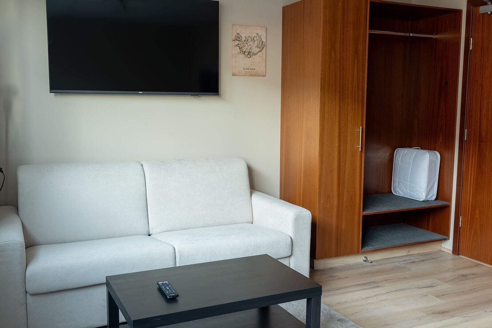 A modern living room with a white sofa, a small black table, a wooden wardrobe, and a flat-screen TV mounted on the wall.