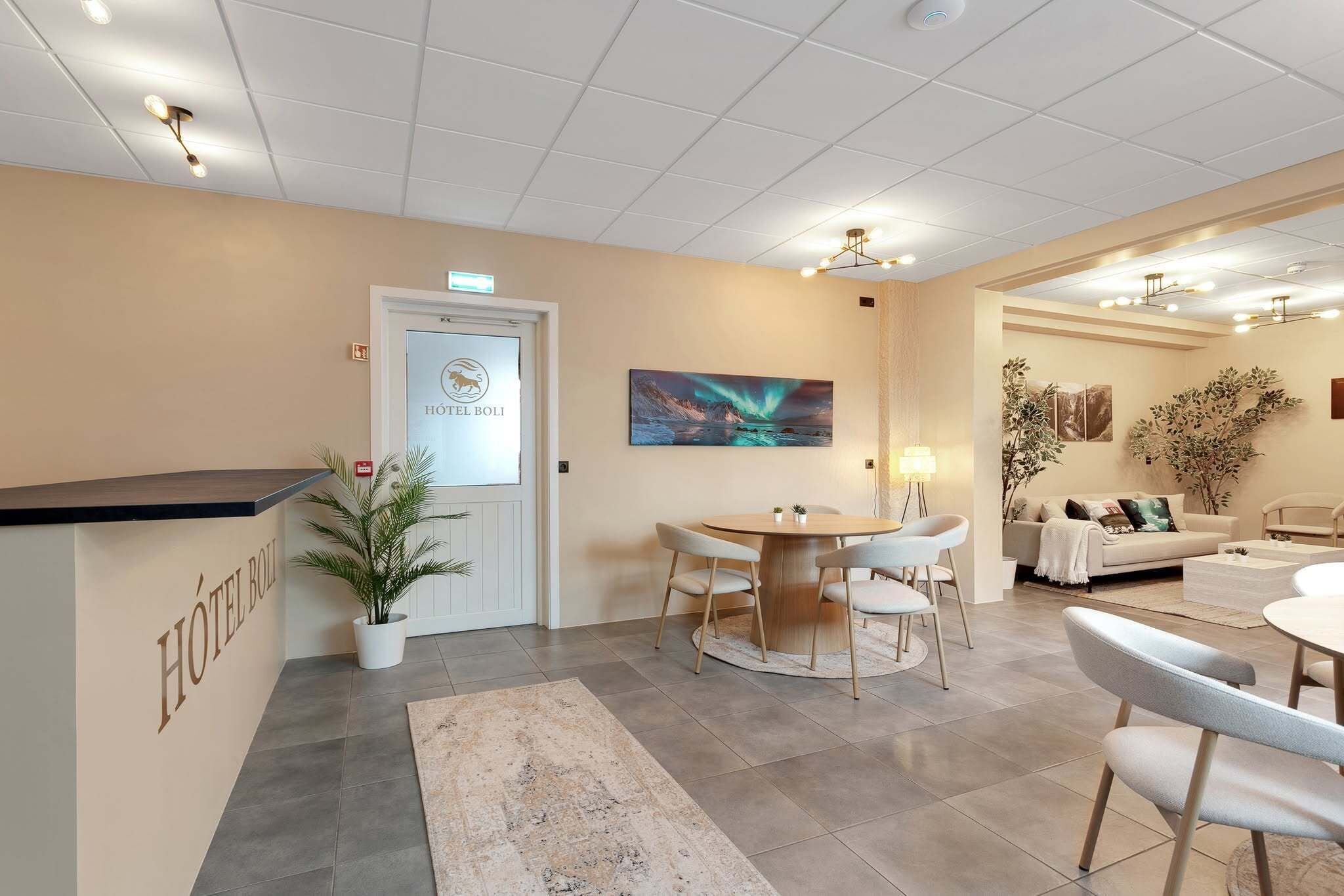 A hotel reception area with a reception desk, a dining area with a round table and chairs, a sofa, and a large window with a view of the night sky.