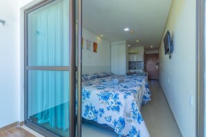 A hotel room with a large sliding glass door leading to a balcony, a bed with floral bedding, a small table, and a TV mounted on the wall. - Manawa Beach #a33 - Porto de Galinhas By Carpediem (Ipojuca)