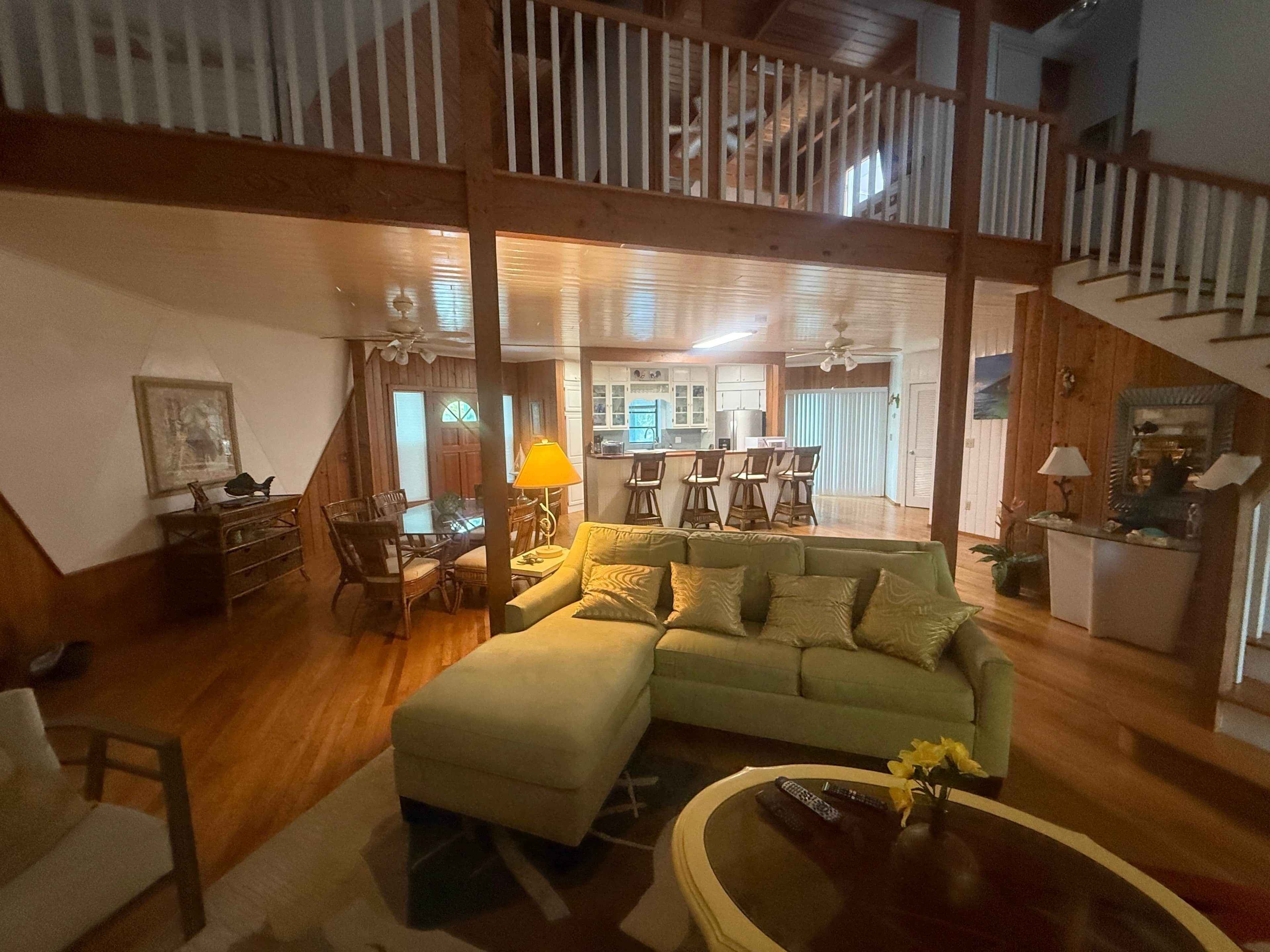 A spacious living area with a large sofa, a dining table with chairs, a kitchen in the background, and a staircase.