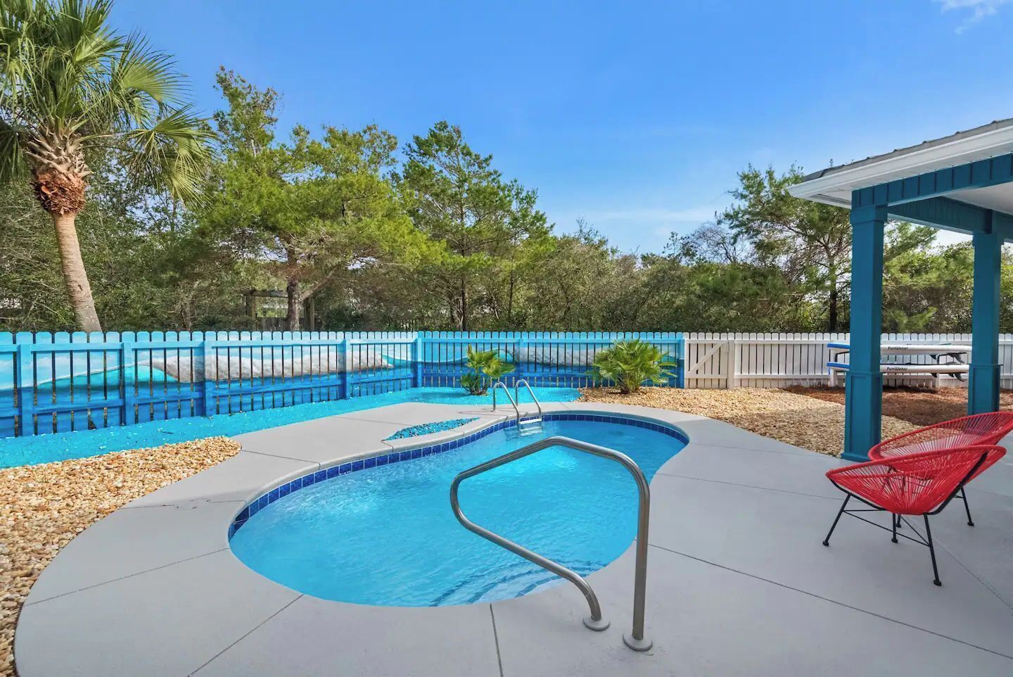 A swimming pool with a blue border, surrounded by a blue fence, and a covered area with red chairs.