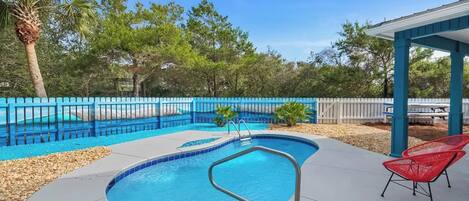 A swimming pool with a blue border, surrounded by a blue fence, and a covered area with red chairs.