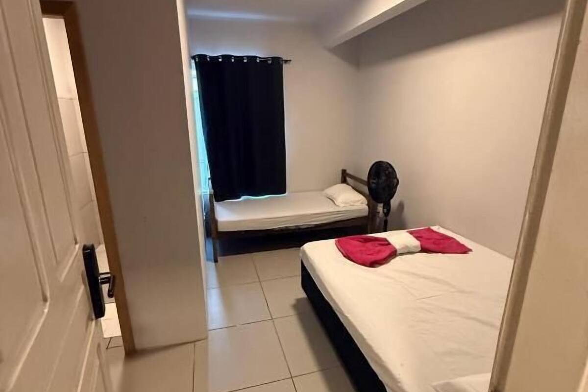 Une petite chambre d’hôtel étroite, avec un lit simple, une table de chevet et une fenêtre avec un rideau.