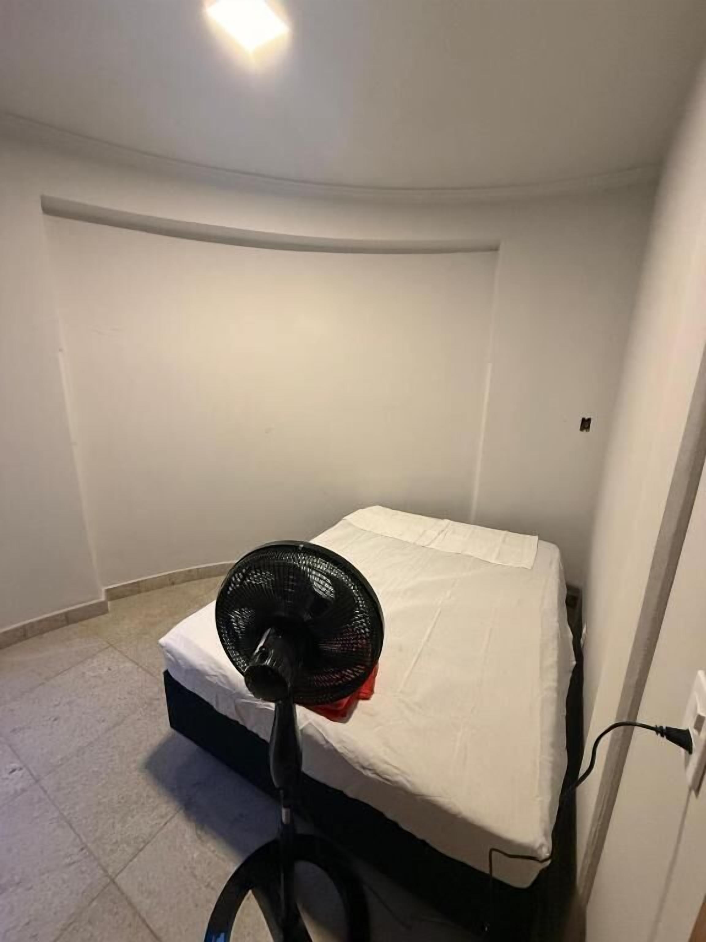 A small, single-bed room with a standing fan in front of the bed.