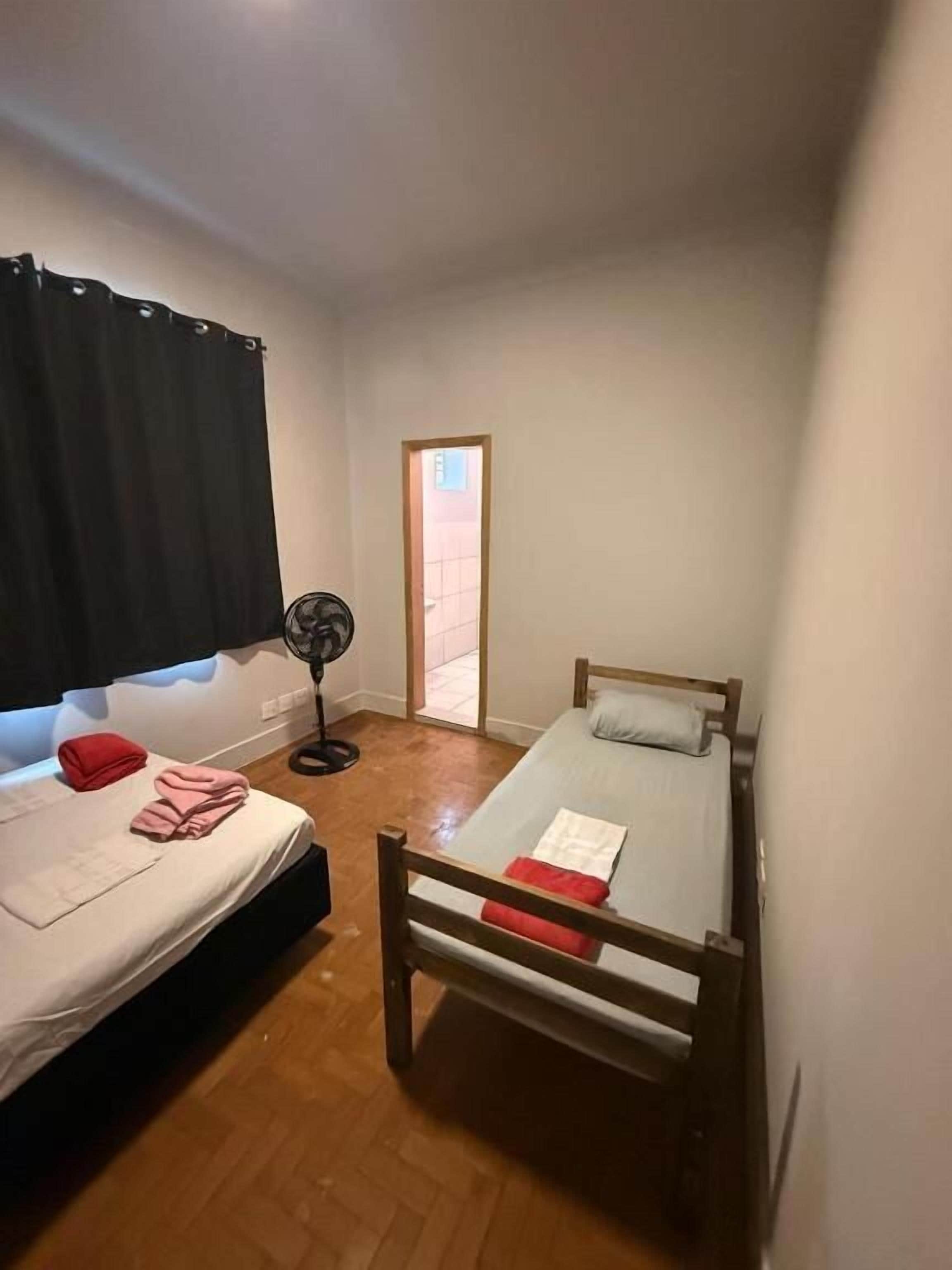 A small, single-room accommodation with two beds, a fan, and a window with curtains.
