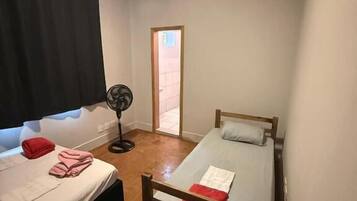 A small, single-room accommodation with two beds, a fan, and a window with curtains.