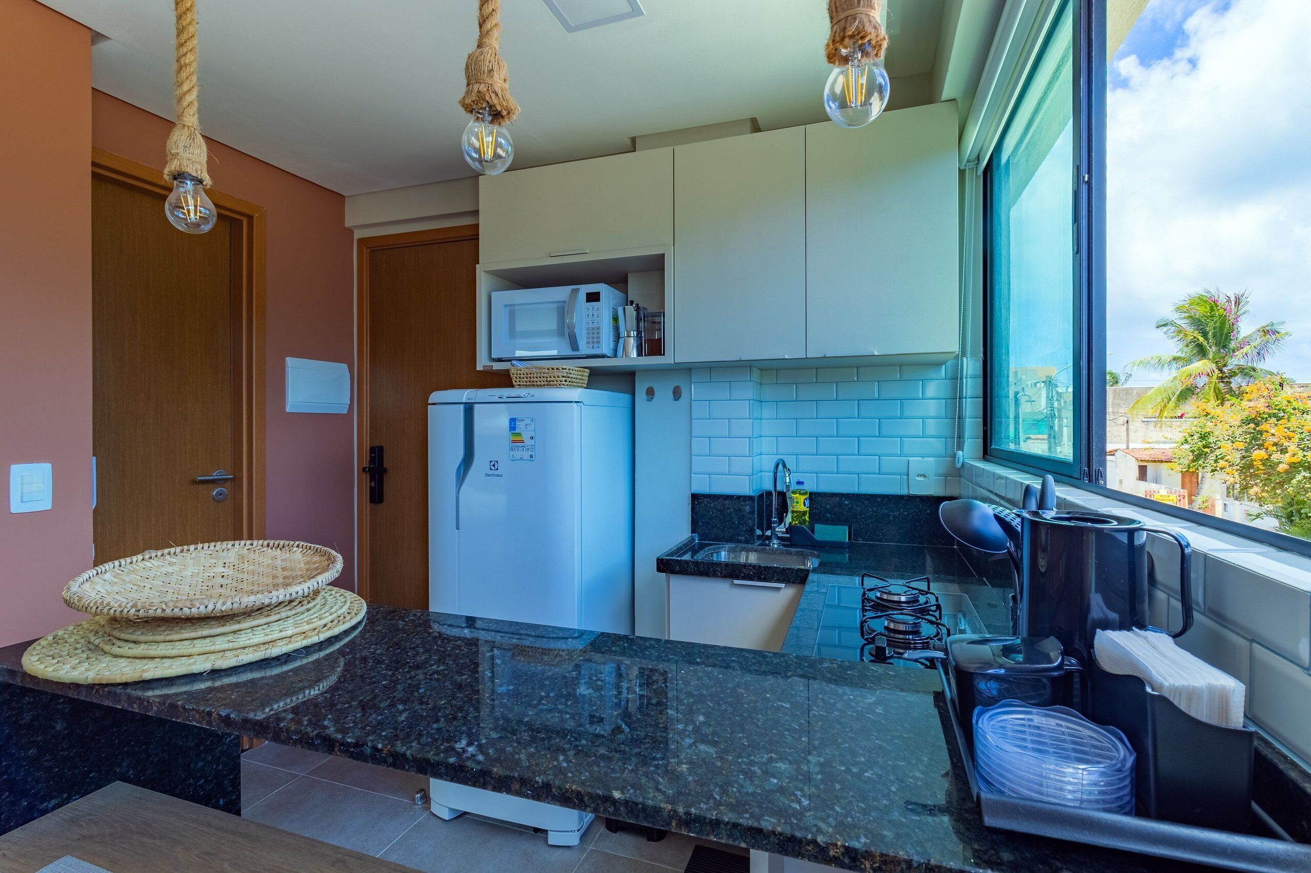A modern kitchen with a granite countertop, white cabinets, a microwave, and a refrigerator. There are hanging lights and a window with a view of palm trees.