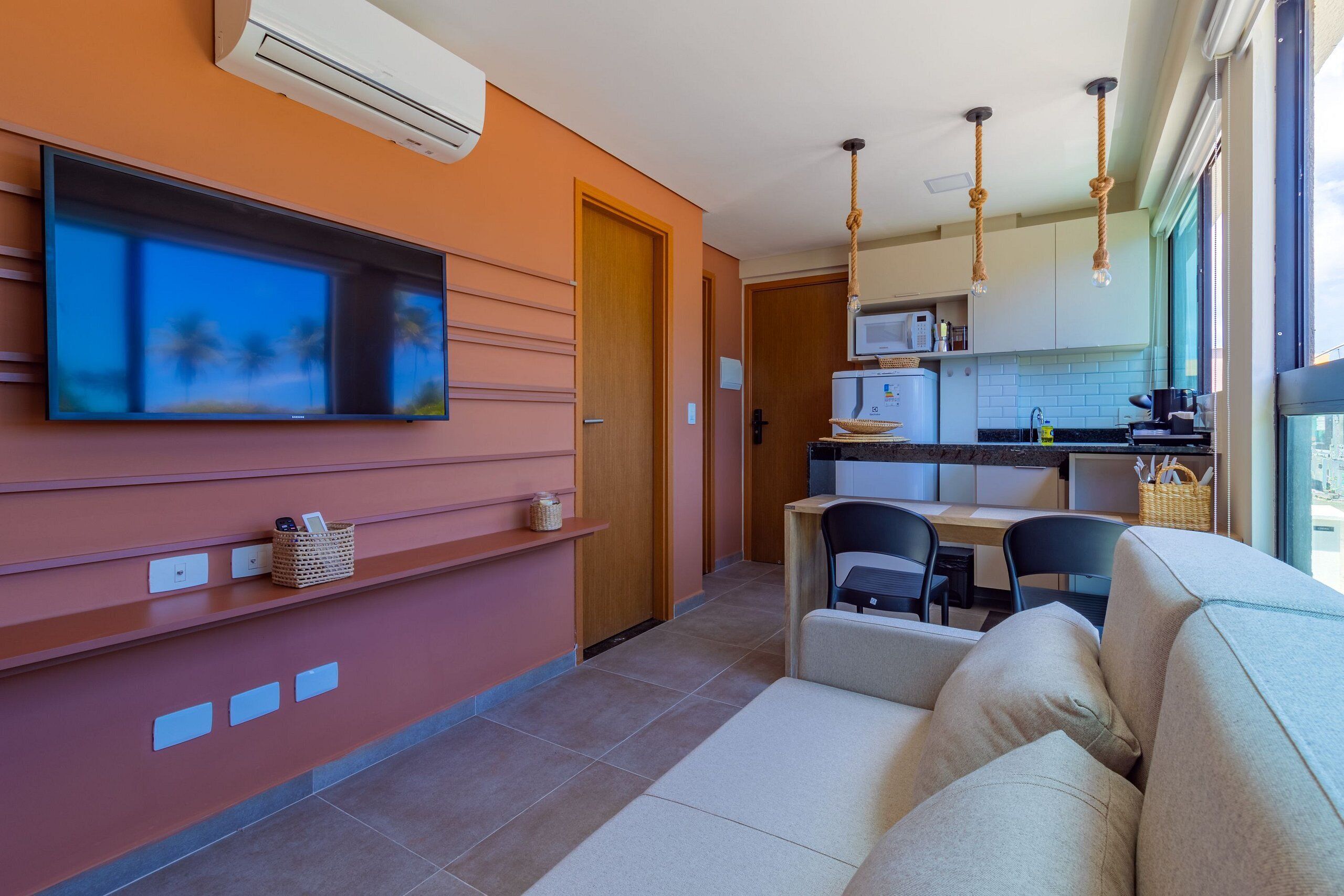 A modern kitchen with a flat-screen TV, a dining area with a sofa, and a large window providing natural light.