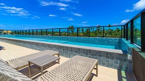 Piscina infinita no terraço com água azul cristalina, rodeada por espreguiçadeiras e um guarda-corpo de vidro.