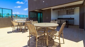 Um terraço no telhado com uma mesa redonda e cadeiras de vime, uma área de cozinha com armários, e vista para o mar.